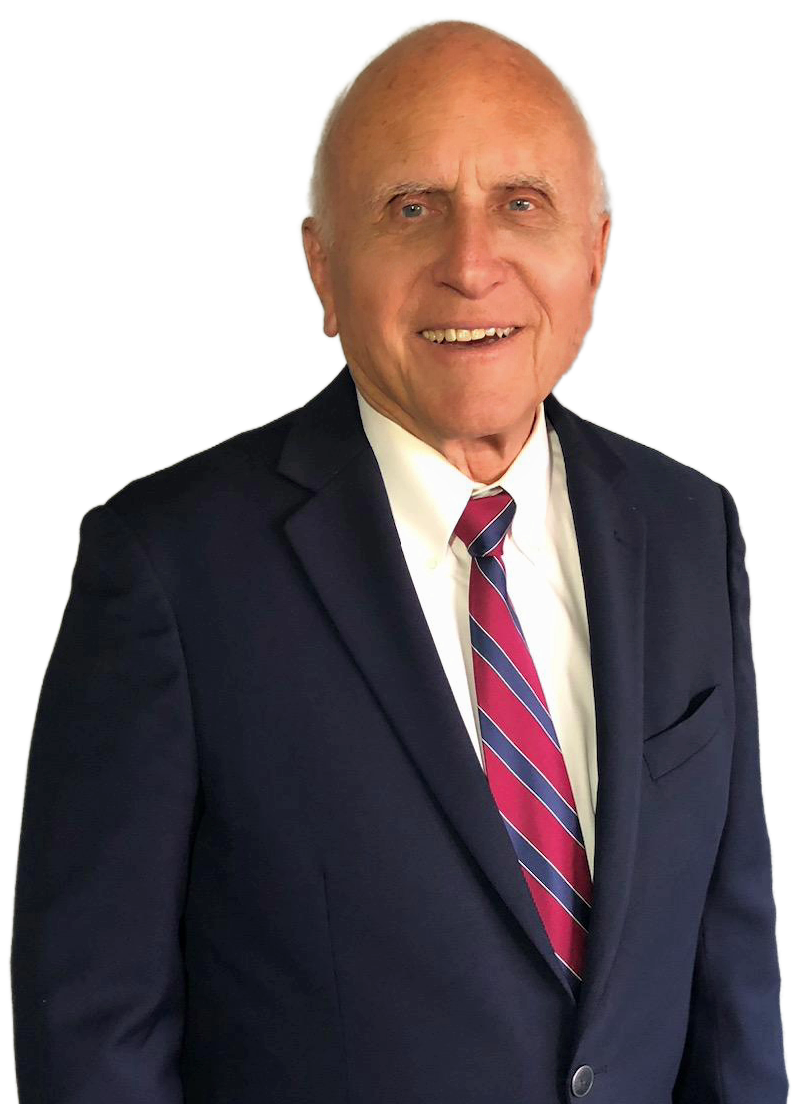 A photograph of an older gentleman with a bald head, light skin, and a warm smile. He is wearing a navy blue suit, a white dress shirt, and a red and blue striped tie.